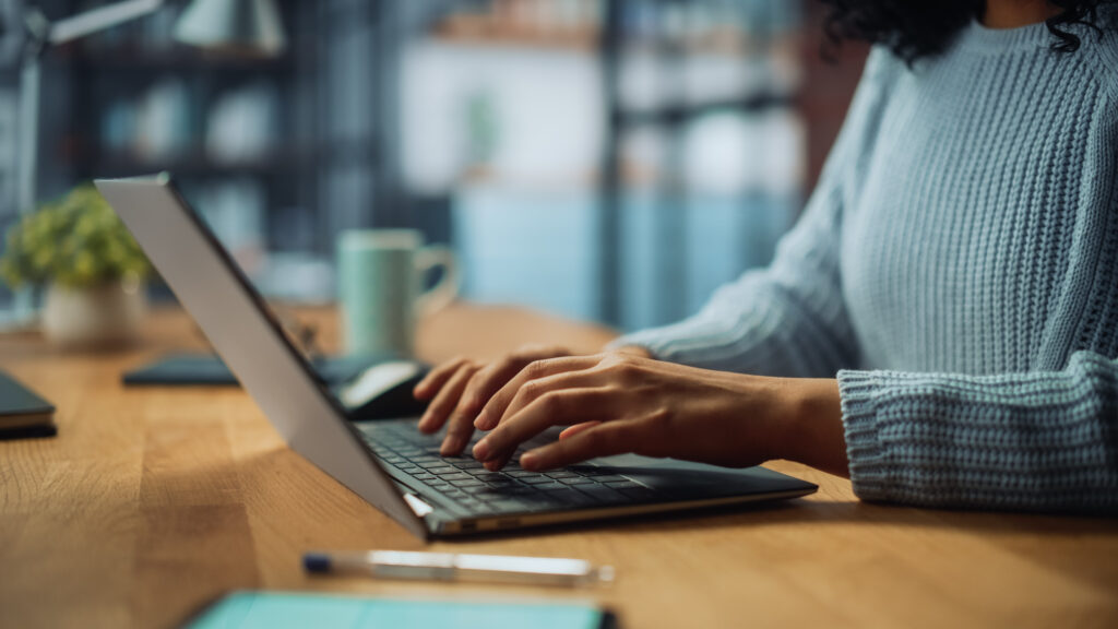 A close-up, side view of a person's hands typing on a laptop keyboard. The person is wearing a light blue ribbed sweater and is seated at a wooden desk in a softly blurred office or home office setting. A pen and notebook are visible in the foreground, with a desk lamp and a mug in the background.
