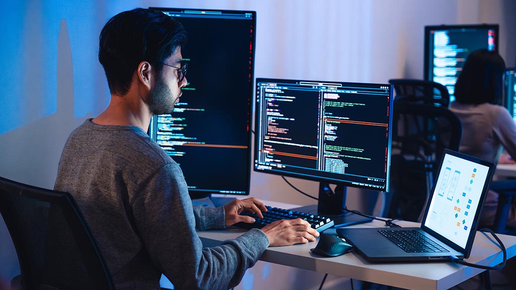 A side view of a programmer wearing glasses and a grey sweater, focused on two large computer monitors and a laptop displaying programming code. The room is lit with blue ambient light, and another person is visible in the background.